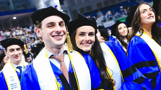 A group of smiling graduates in blue robes and caps with yellow tassels and white stoles stand together at a graduation ceremony, surrounded by a cheering crowd.
