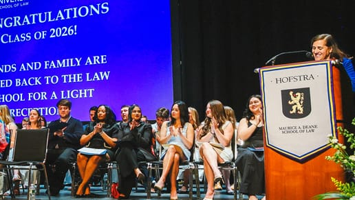 A group of people sit and clap on stage at a Hofstra law school event, while a woman speaks at a podium displaying the Hofstra logo and a large screen congratulates the Class of 2026.