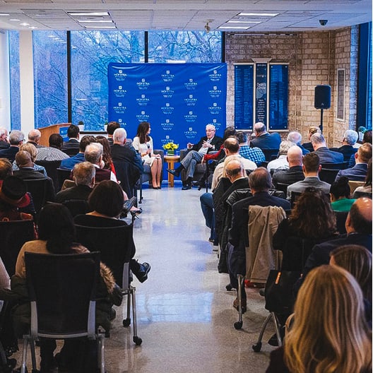 An audience sits facing a stage where two people are seated and conversing in front of a blue backdrop with white logos, in a room with large windows and brick walls.