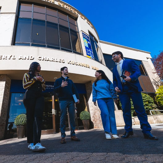 Four people stand and talk outside a building with large windows and a curved sign that reads Sheryl and Charles Kushner Hall. The group appears to be students, carrying bags and books, under a clear blue sky.