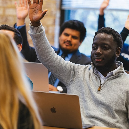 A man sitting at a desk with a laptop raises his hand in a classroom setting. Other students are also visible, some with their hands raised, suggesting active participation.
