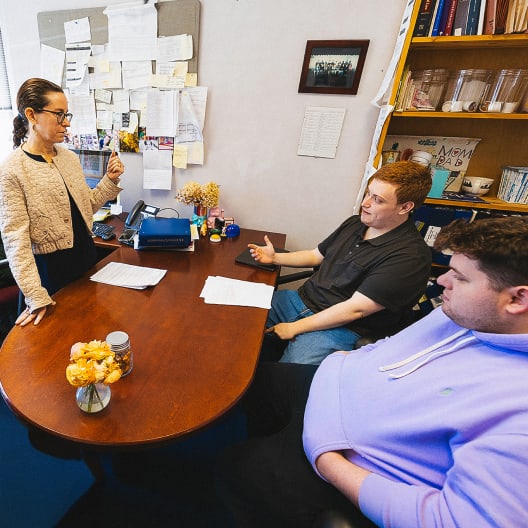 A woman stands and speaks to two seated young men in an office. Papers, flowers, and a typewriter are on the desk, with shelves of books and a bulletin board in the background.