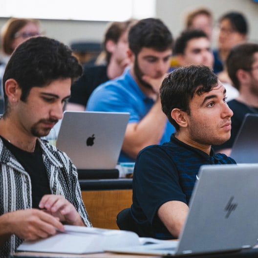 Several students sit in a classroom, focused on their laptops and notes. Some are writing or reading, while others listen attentively. The scene suggests an engaged learning environment.
