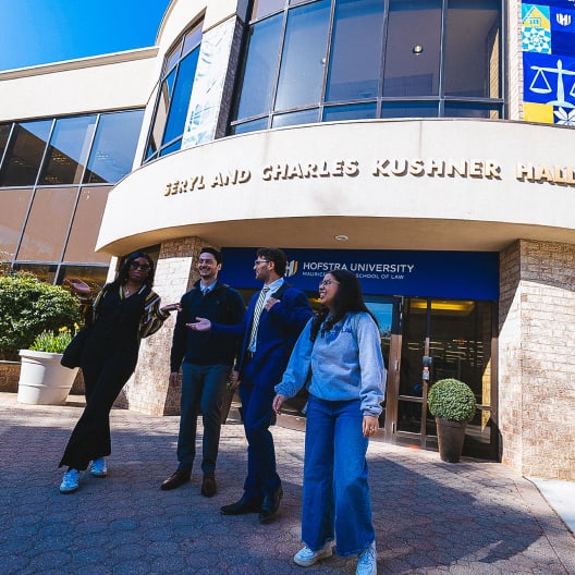 Four people stand and talk outside Seryl and Charles Kushner Hall at Hofstra University School of Law, with the building entrance and signs visible behind them on a sunny day.