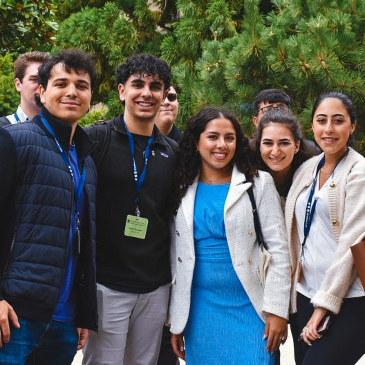 A group of young adults wearing conference badges stand together smiling outdoors, with green trees in the background. The people are dressed in business casual attire.