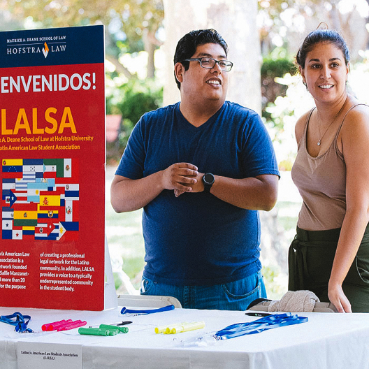 Two people stand behind a table with pens and lanyards, next to a colorful LALSA welcome sign for the Latin American Law Student Association at Hofstra Law. The setting appears to be an outdoor event.