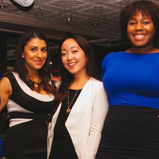 Three women smiling and posing together indoors at what appears to be a social or professional event. One wears a black and white dress, one a white jacket, and one a bright blue dress.