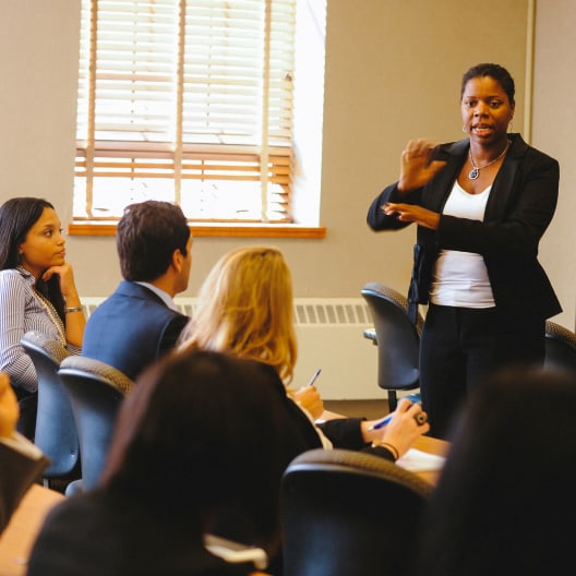 A woman in business attire stands and speaks to a group of seated people in a conference room, gesturing with her hands while others listen and take notes.