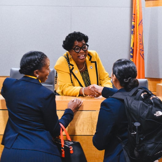 A woman in a yellow blazer smiles and shakes hands with another woman in a suit, while a third woman stands nearby. They are in a formal indoor setting with a flag in the background.