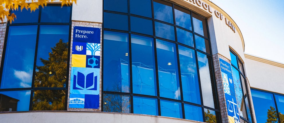 A modern building with large curved glass windows, blue banners displaying abstract designs and the words Prepare Here, and part of the words School of Law visible at the top. Trees are reflected in the windows.