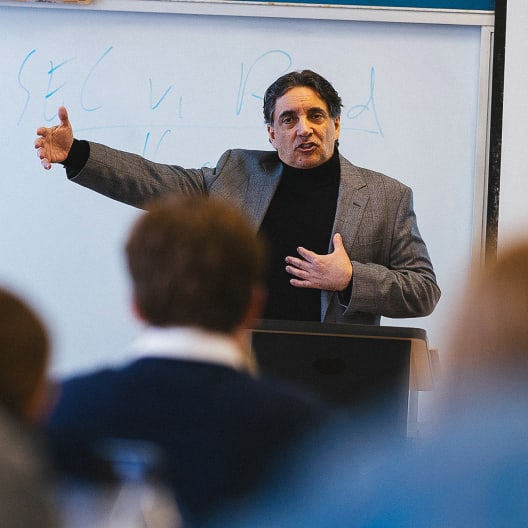 A man in a gray blazer and black turtleneck gestures while speaking in front of a whiteboard to an audience. Writing is visible on the board behind him.