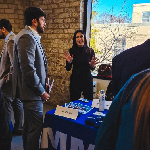 A woman enthusiastically talks to a man in a suit at a career fair booth by a window, with promotional items and papers on a blue table. Other attendees are visible nearby.