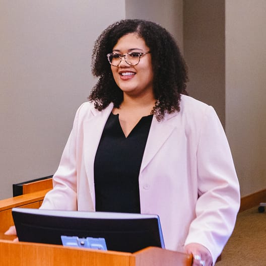A woman with curly hair and glasses, wearing a light pink blazer and black top, stands at a podium with a monitor, smiling as she speaks in a classroom or conference setting.