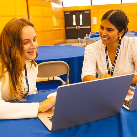 Two women sit at a table with blue tablecloths, smiling and working together on laptops in a bright room with wood-paneled walls and large windows in the background.