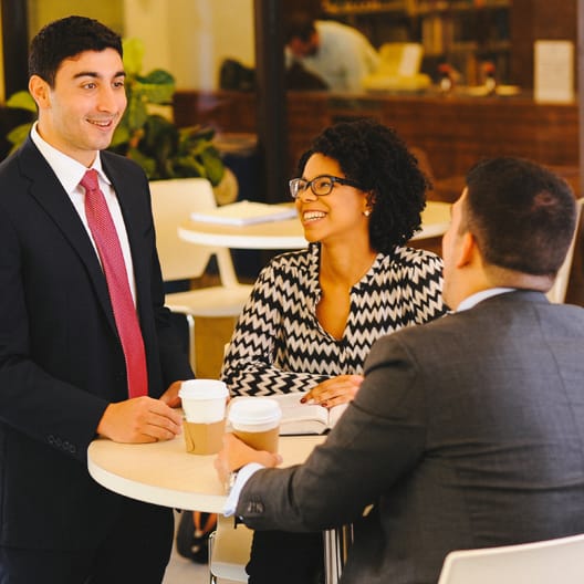 Three professionally dressed people converse around a small table in an office setting, with two seated and holding coffee cups, and one person standing and smiling.