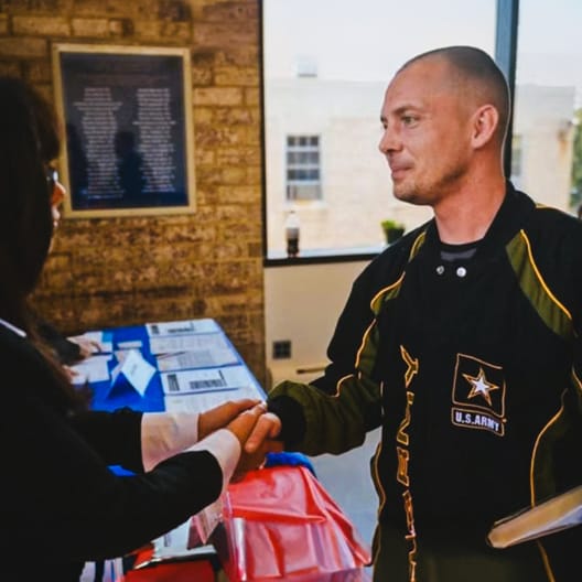 A man in a U.S. Army uniform shakes hands with a woman across a table indoors, with papers and a plaque visible in the background.