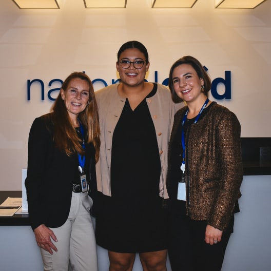 Three women stand together smiling in front of a reception desk with a partially visible company sign on the wall behind them. All are dressed in business attire and wearing blue lanyards.