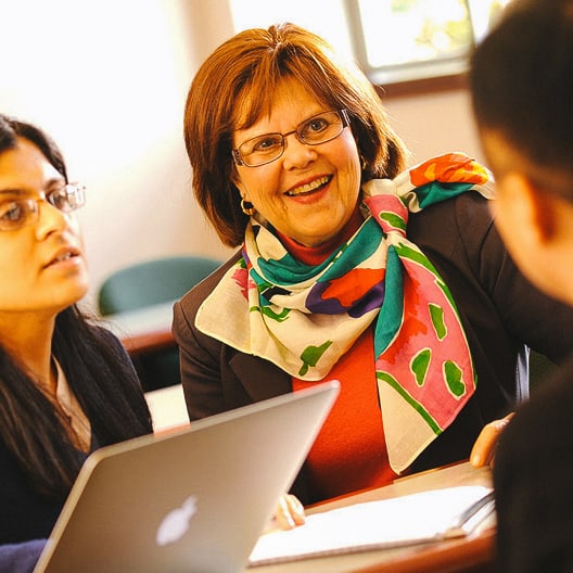 Three people sit at a table having a discussion. A woman in a colorful scarf is smiling, another woman with glasses is looking ahead, and a third person is partially visible. A laptop and documents are on the table.