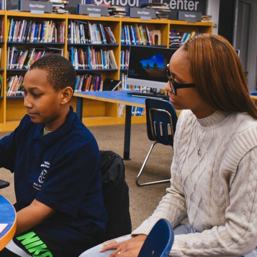 A young boy and a woman sit together at a table in a library. The boy works on a tablet while the woman, wearing glasses and a cream sweater, attentively watches him. Bookshelves and computer monitors are in the background.