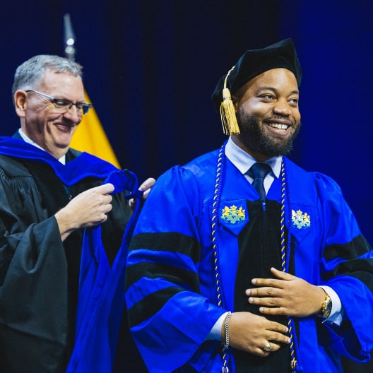 A smiling graduate in a blue academic robe and cap is hooded by a faculty member on stage during a graduation ceremony. Both appear happy and celebratory, with a dark blue background and flag behind them.