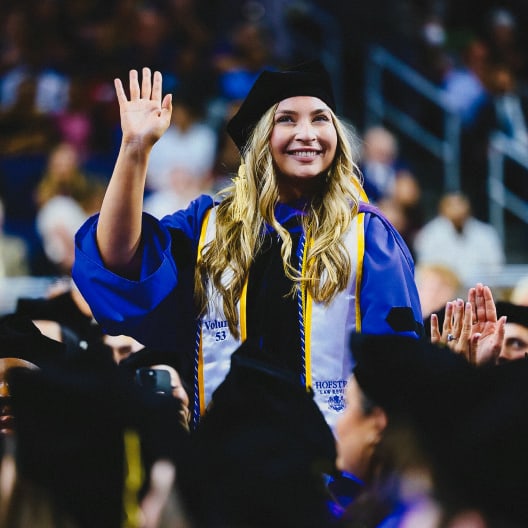 A smiling graduate in a cap and gown waves while standing among a crowd of graduates at a graduation ceremony.