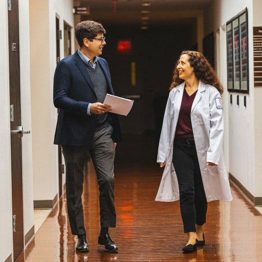 A man in business attire and a woman in a white lab coat walk and talk in a hallway with wooden floors, both smiling at each other.