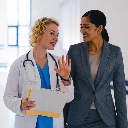 A doctor holding a clipboard and greeting a woman in a business suit as they walk together in a bright hallway, both smiling and engaged in conversation.