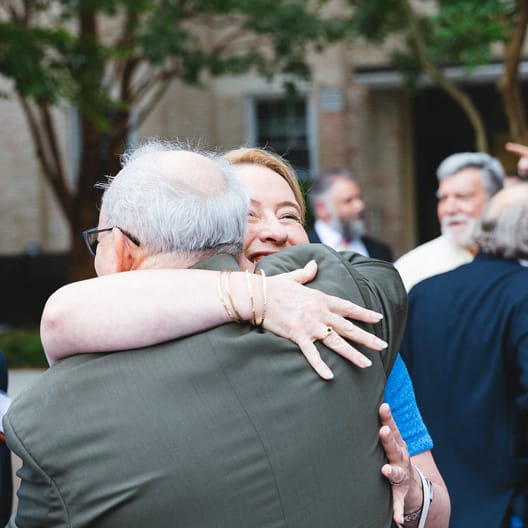 A woman smiling brightly as she hugs an older man outdoors, surrounded by other people in conversation. Trees and buildings are visible in the background.
