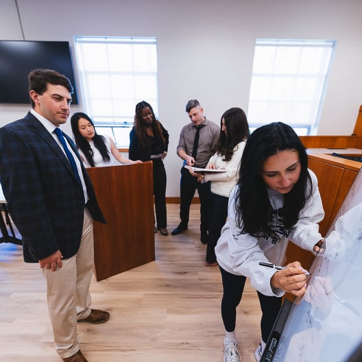 A group of students and a man in a suit stand in a classroom. One student writes on a whiteboard while others discuss and observe in the background. Sunlight streams through two large windows.