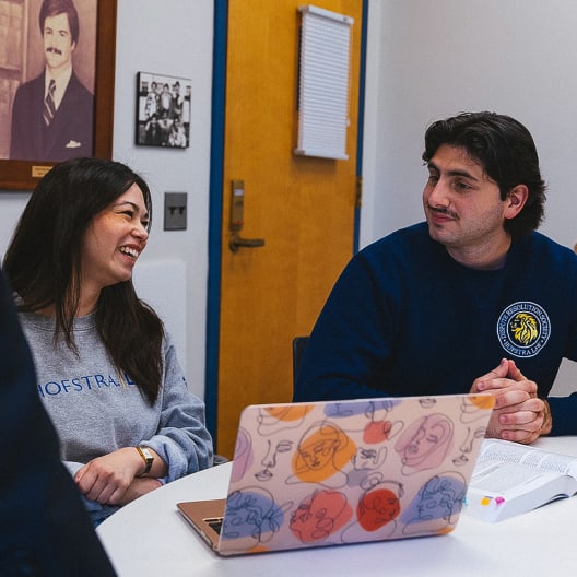 Two students sit at a table in a classroom, talking and smiling. One has a colorful laptop, and there are textbooks on the table. A framed portrait and a photo are on the wall behind them.