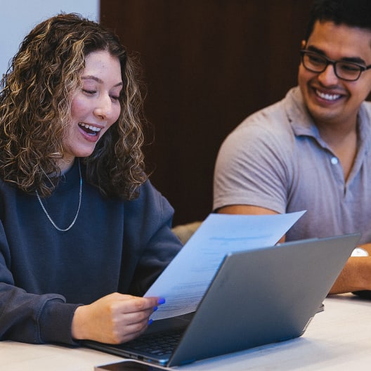 A young woman smiling while holding a sheet of paper, sitting beside a young man who is also smiling. They are at a table with an open laptop in front of them.