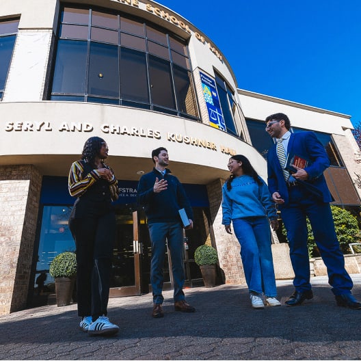 Four people stand and talk outside a modern building with large windows and a sign reading Seryl and Charles Kushner Hall under a bright blue sky. Two hold bags and one carries a laptop.