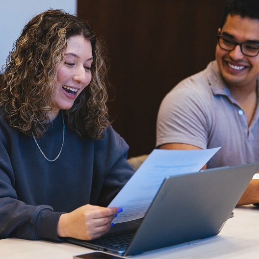 Two people sit at a table with laptops, smiling and holding papers. The woman on the left looks excited while reading her paper, and the man on the right is smiling and looking at her.