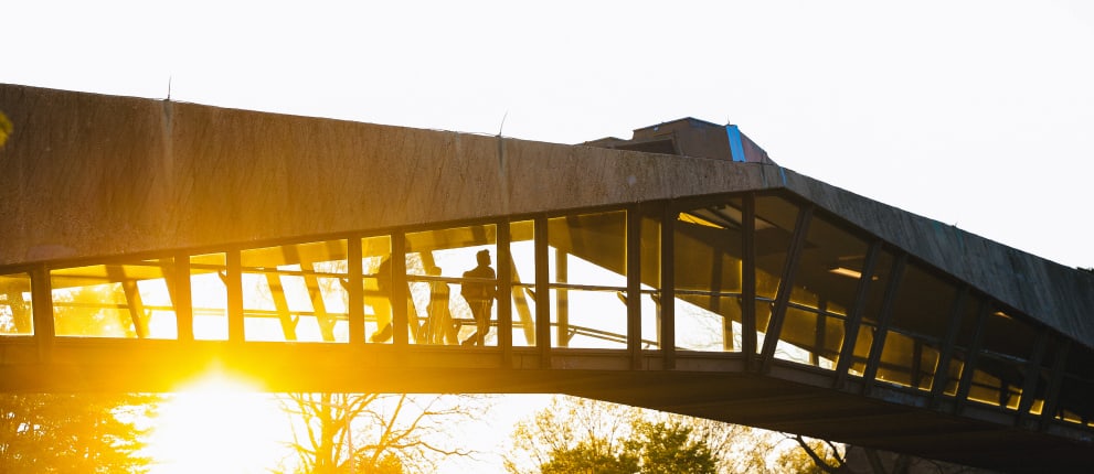 Silhouettes of people walking inside a glass-walled, concrete bridge or walkway at sunset, with bright sunlight streaming through the windows and illuminating the structure.