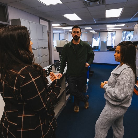Three people stand together in an office, having a conversation. One man leans against a shelf while two women face him, engaged in discussion. The office has blue carpet and large windows in the background.