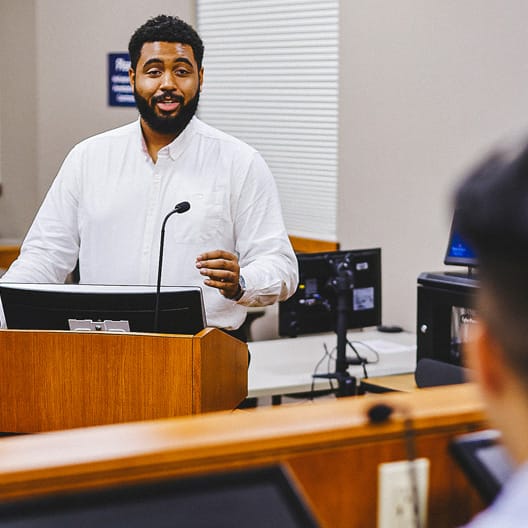 A man with a beard, wearing a white shirt, stands at a podium speaking to an audience in a classroom or lecture hall, with a computer monitor and microphone in front of him.