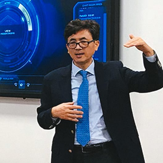 A man in a suit and blue tie gestures with one hand raised while speaking in front of a digital display showing circular blue graphics.