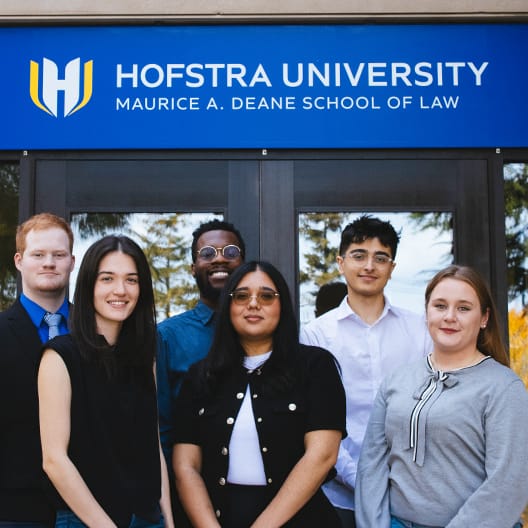 Six young adults stand in front of the Hofstra University Maurice A. Deane School of Law sign, posing and smiling for the photo.