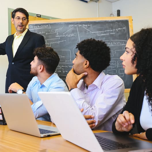 A teacher stands by a chalkboard covered with notes and diagrams, speaking to three students seated at a table with open laptops, attentively listening in a classroom setting.