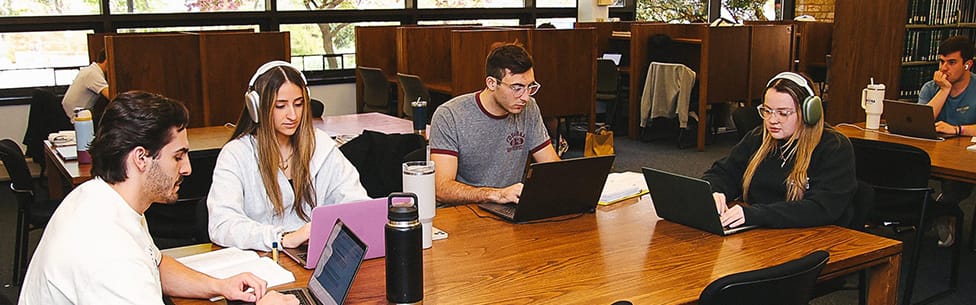 Four students sit at a wooden table in a library or study area, each working on laptops. Three of them wear headphones. Books, notebooks, and water bottles are on the table. Other students work in the background.