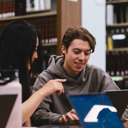 Two people sitting at a table in a library, smiling and looking at a laptop screen together, with bookshelves in the background and another laptop visible in the foreground.