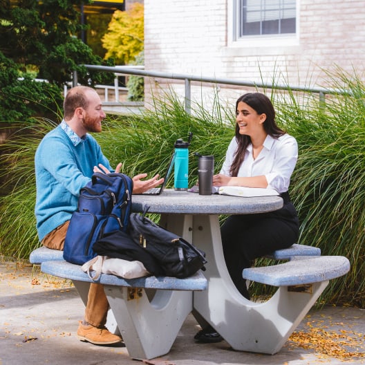 Two people sit at an outdoor concrete table, talking and smiling. Both have backpacks and drinks on the table. Green grass and a building are in the background. It appears to be a pleasant, casual meeting.