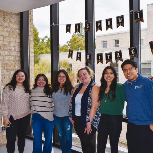 Six people stand smiling in front of large windows with a banner reading Hispanic Heritage. They are casually dressed and appear to be celebrating in a bright indoor space. Trees and buildings are visible outside.