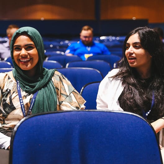 Two women sit and smile while talking in a mostly empty auditorium. One woman wears a green hijab and patterned shirt, and the other has long dark hair and a white top. Blue chairs and blurred people are in the background.
