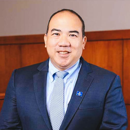 Professor Julian Ku in a dark suit, light blue shirt, and patterned tie smiles at the camera. He is standing in front of a wooden panel wall and is wearing a small blue pin on his lapel.