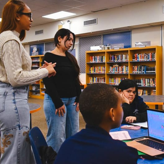 Four students are gathered around a table in a library, engaged in discussion. Two are standing, while two are seated near an open laptop. Shelves filled with books are visible in the background.
