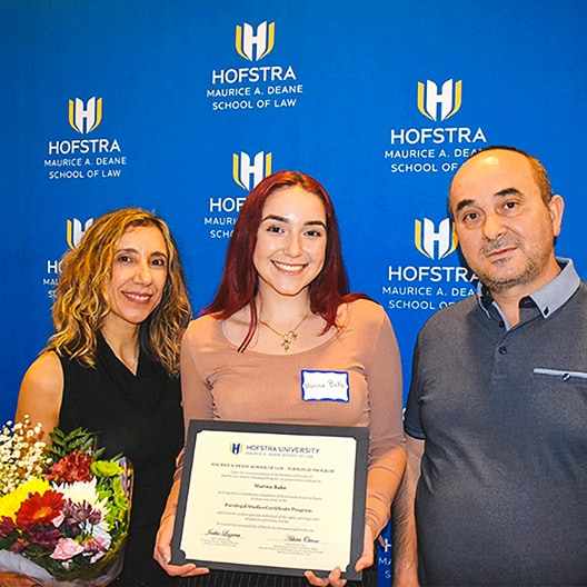 Three people pose in front of a Hofstra Law backdrop. The center person holds a certificate and wears a name tag. The person on the left holds a bouquet of flowers. All are smiling at the camera.