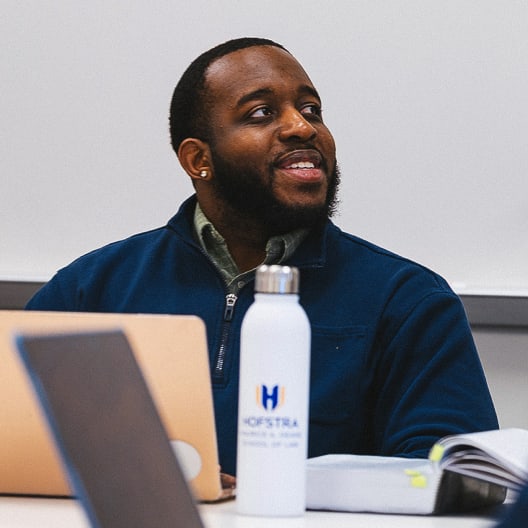 A person sits at a table with a laptop, a water bottle labeled Hofstra, and an open book, smiling and looking to the side in a classroom or meeting setting.
