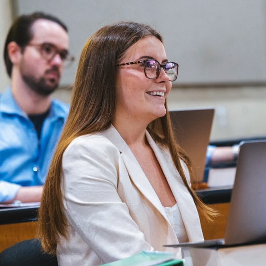 A woman with long brown hair and glasses smiles while sitting at a desk with a laptop in a classroom. A man in a blue shirt is seated behind her, also looking forward.