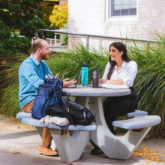 Two people sit at an outdoor concrete picnic table, talking and smiling. Both have backpacks and drinks on the table. The scene is set in a campus-like area with green plants and a building in the background.